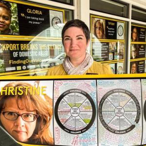 Julia Pierce, Deputy Director of the Rockport Public Library, stands in front of one of the 17 posters of survivors in the Finding Our Voices window exhibit that runs during October’s Domestic Abuse Awareness Month, and that can be viewed by standing outside. (Photo by Patrisha McLean)