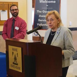 Governor Janet Mills speaks during 2023 award ceremony in the Hall of Flags while Volunteer Maine commissioner Luke Shorty looks on. (Photo courtesy Volunteer Maine)